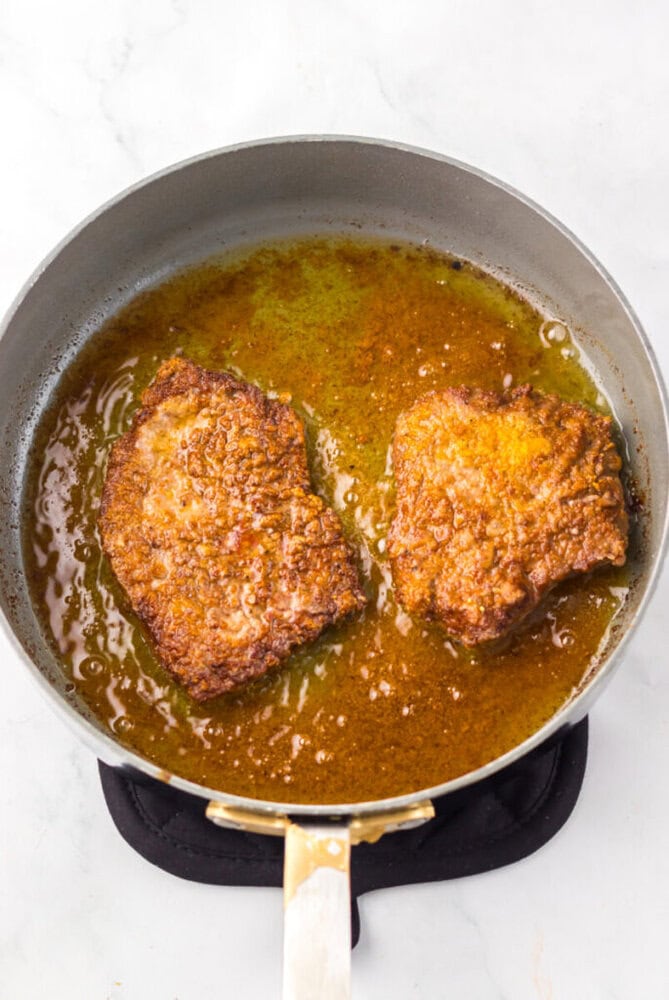Two pieces of fried chicken are cooking in a skillet with oil and seasonings on a stovetop.