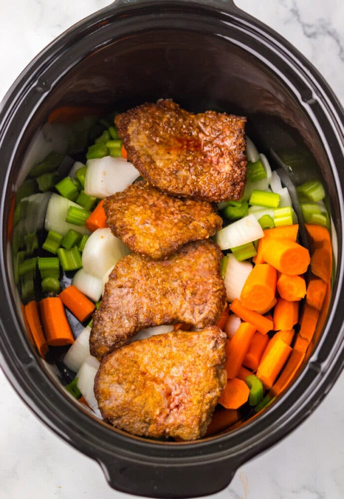 Four breaded beef pieces placed over chopped carrots, celery, and onions inside a slow cooker.