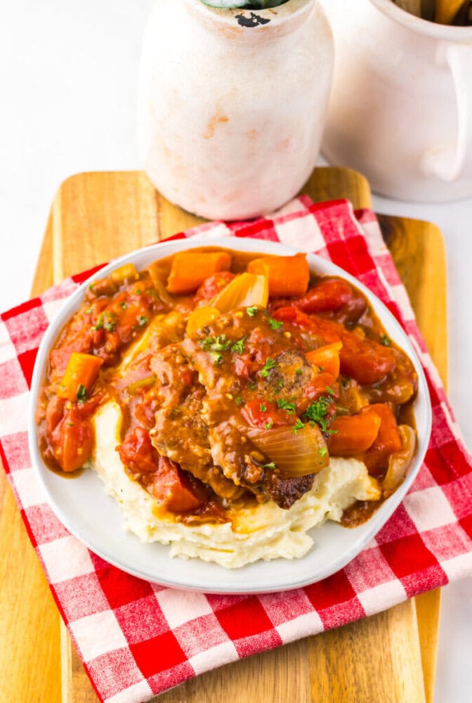 A plate of mashed potatoes topped with a beef and vegetable stew, placed on a red and white checkered napkin on a wooden board.