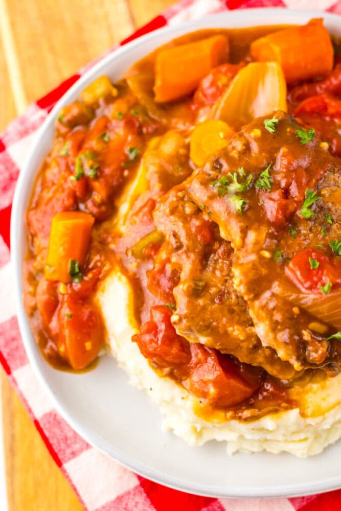 A plate of mashed potatoes topped with beef, carrots, and tomatoes in a brown gravy, served on a white plate with a red plaid napkin underneath.