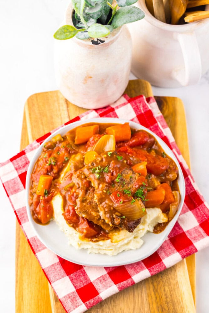 A plate of Salisbury steak with tomato-based gravy, carrots, and onions served over mashed potatoes on a wooden board with a red and white checkered napkin.