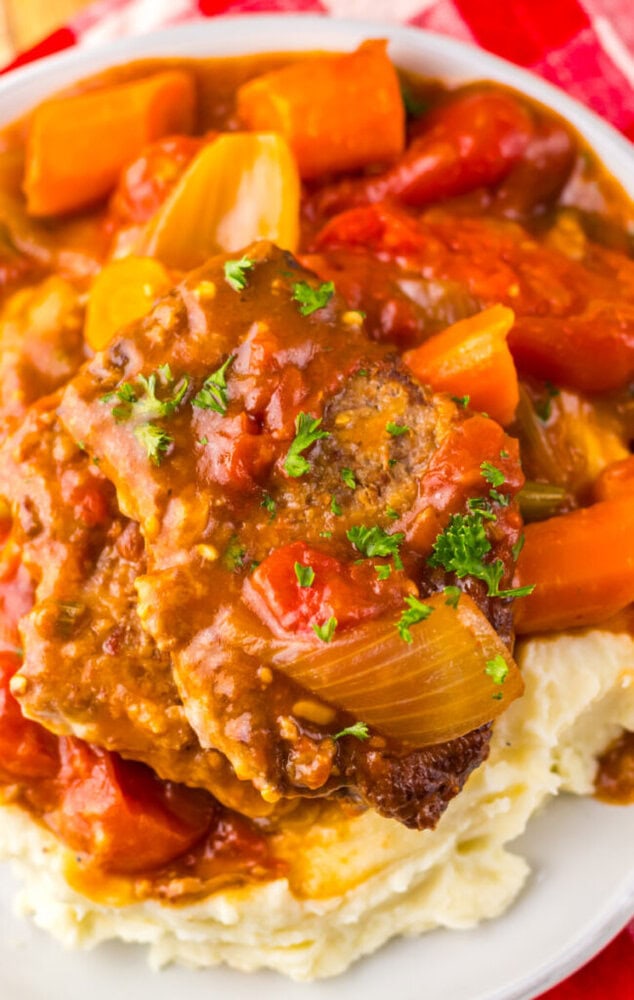 A plate of mashed potatoes topped with a slice of meatloaf, chunky tomato-based sauce, carrots, onions, and garnished with chopped parsley.