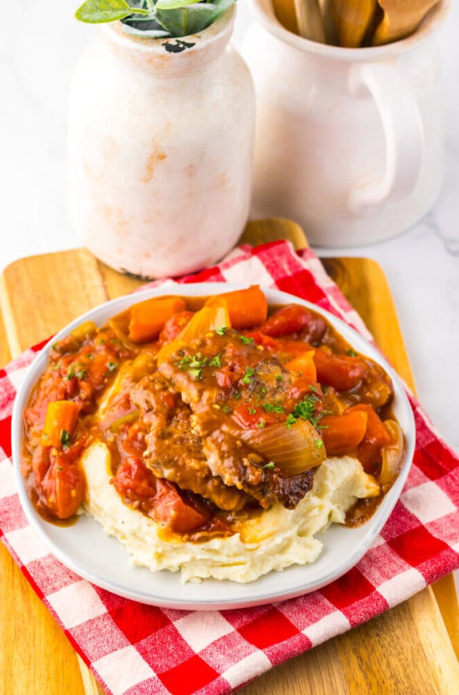 A plate of mashed potatoes topped with beef, carrots, and tomato-based gravy sits on a red and white checkered napkin, with a white jar in the background.