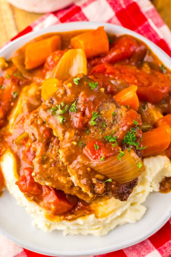 A plate of mashed potatoes topped with braised beef, carrots, onions, and tomato-based gravy, garnished with chopped parsley.