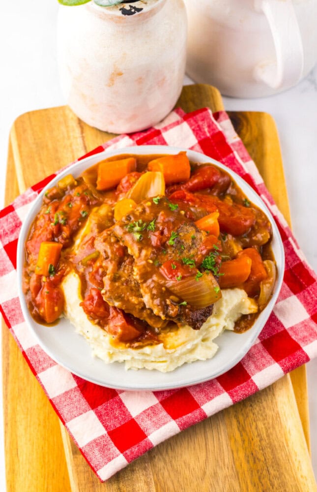 A plate of mashed potatoes topped with beef stew, carrots, and tomatoes, garnished with parsley, is set on a red and white checkered napkin on a wooden board.