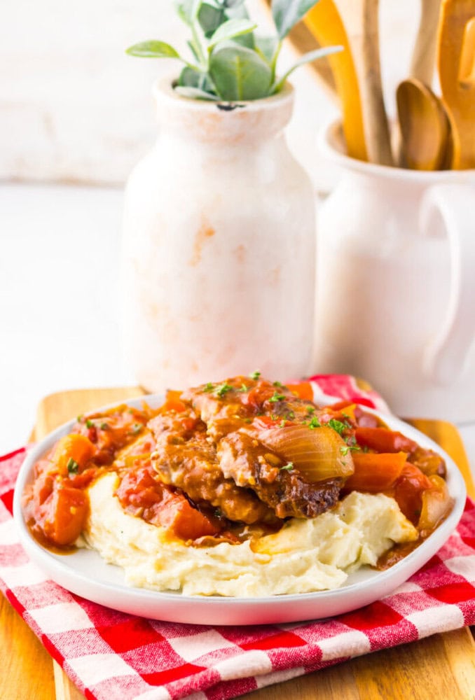 A plate of mashed potatoes topped with smothered chicken and vegetables, set on a red-checkered cloth with a vase and utensils in the background.