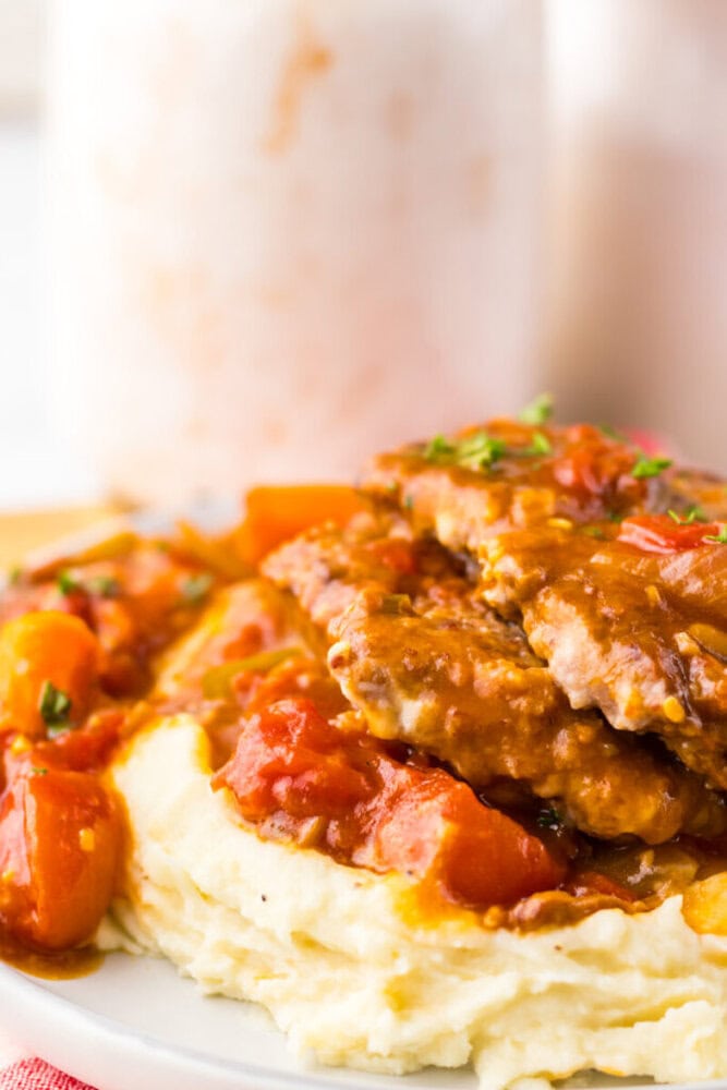 A plate of mashed potatoes topped with pieces of meat in tomato-based gravy, garnished with herbs.