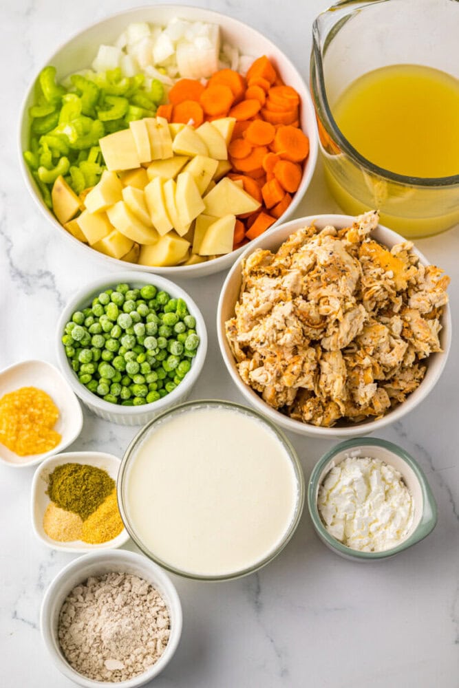 An assortment of ingredients in bowls and a measuring cup, including chopped vegetables, peas, cooked chicken, broth, cream, seasonings, and flour, arranged on a marble surface.