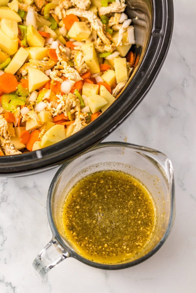 Chopped vegetables and shredded chicken in a slow cooker next to a glass measuring cup filled with seasoned broth on a marble countertop.