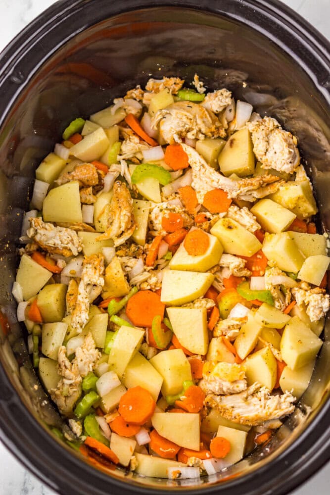 Chopped potatoes, carrots, celery, onions, and shredded chicken inside a slow cooker, prepared for cooking.