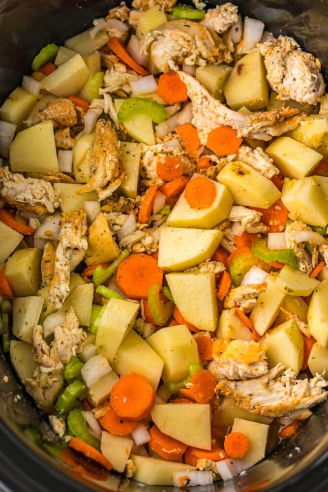 Chopped potatoes, carrots, celery, onions, and shredded chicken in a slow cooker, seasoned and ready to cook.