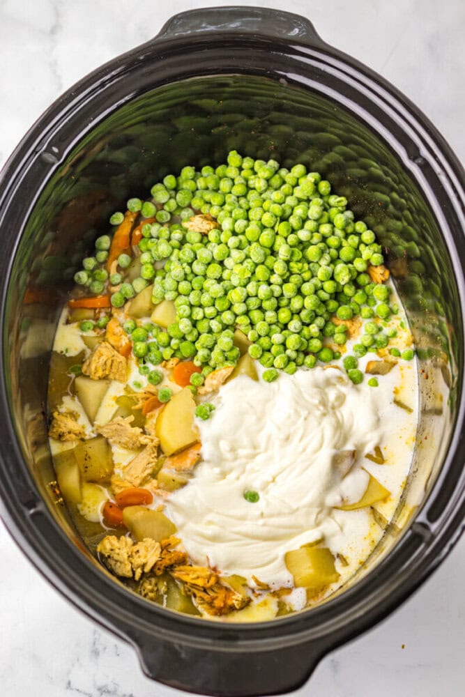 A slow cooker filled with uncooked soup ingredients, including peas, potatoes, carrots, chicken, creamy sauce, and broth.