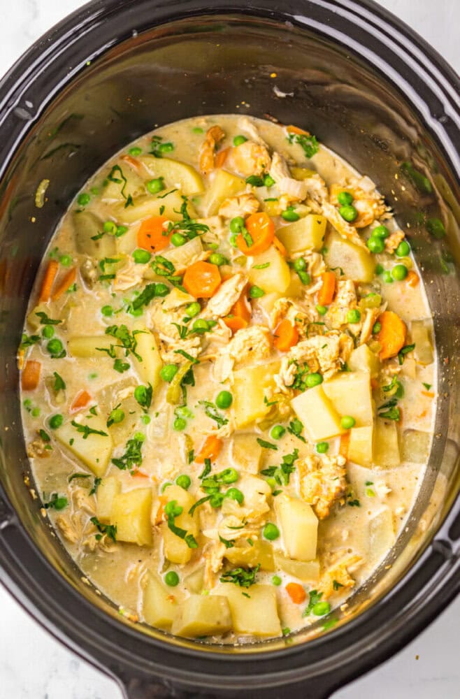A slow cooker filled with a creamy stew containing diced potatoes, carrots, peas, shredded chicken, and chopped herbs.