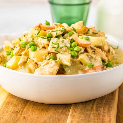 A white bowl filled with a creamy mixture of chicken, peas, carrots, and potatoes sits on a wooden board next to a wooden spoon.