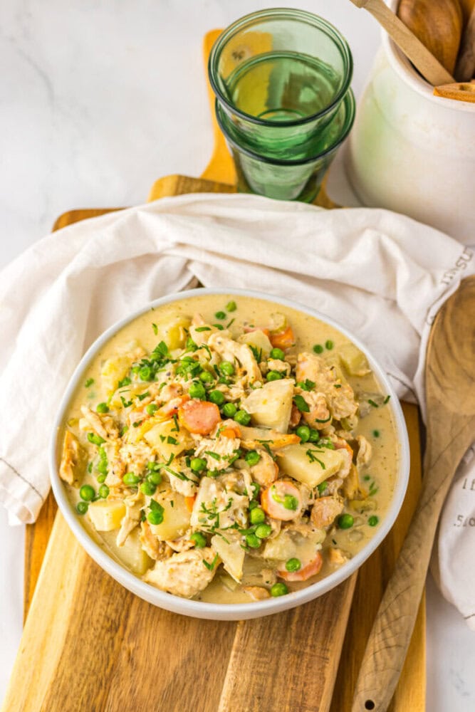 A bowl of creamy chicken stew with potatoes, carrots, and peas sits on a wooden board, next to stacked green glasses, a wooden spoon, and a white cloth.