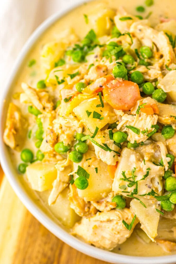 A close-up of a bowl filled with creamy chicken stew, featuring chunks of potato, sliced carrots, green peas, and chopped herbs.
