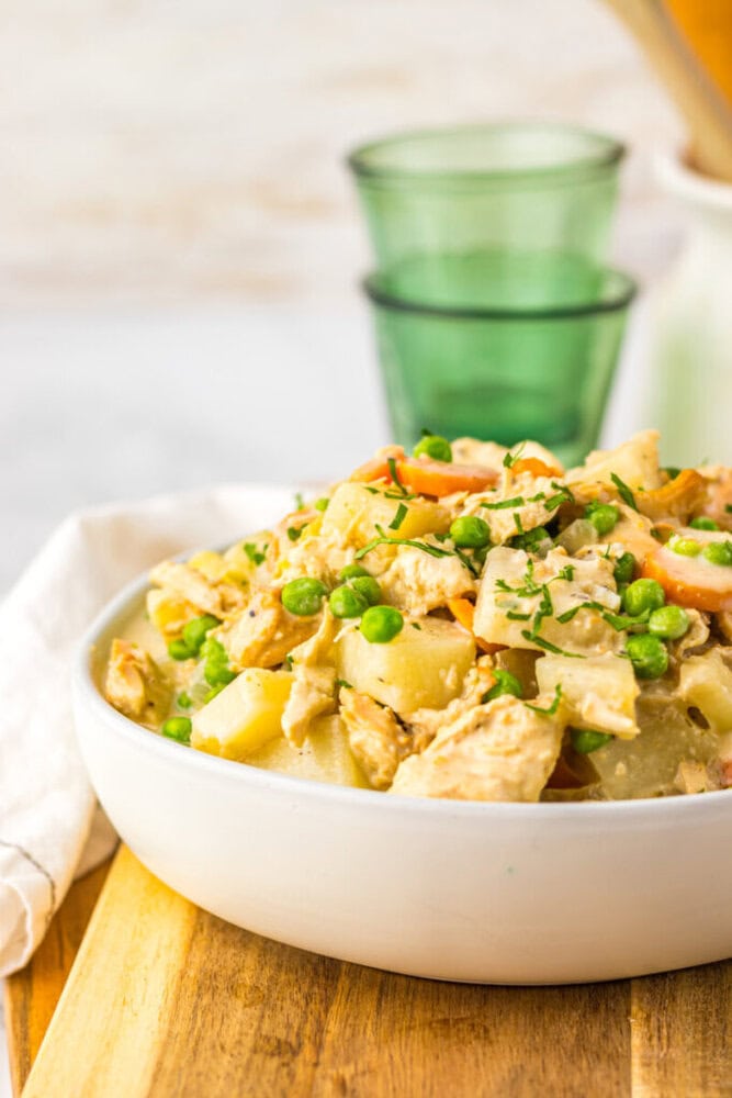 A bowl of chicken salad with potatoes, peas, and chopped herbs on a wooden board, with two green glasses in the background.