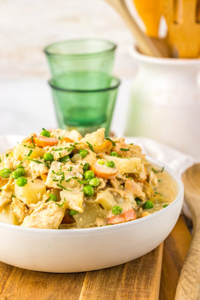 A bowl of creamy chicken and vegetable stew with peas, carrots, and potatoes sits on a wooden board, with green glasses and kitchen utensils in the background.