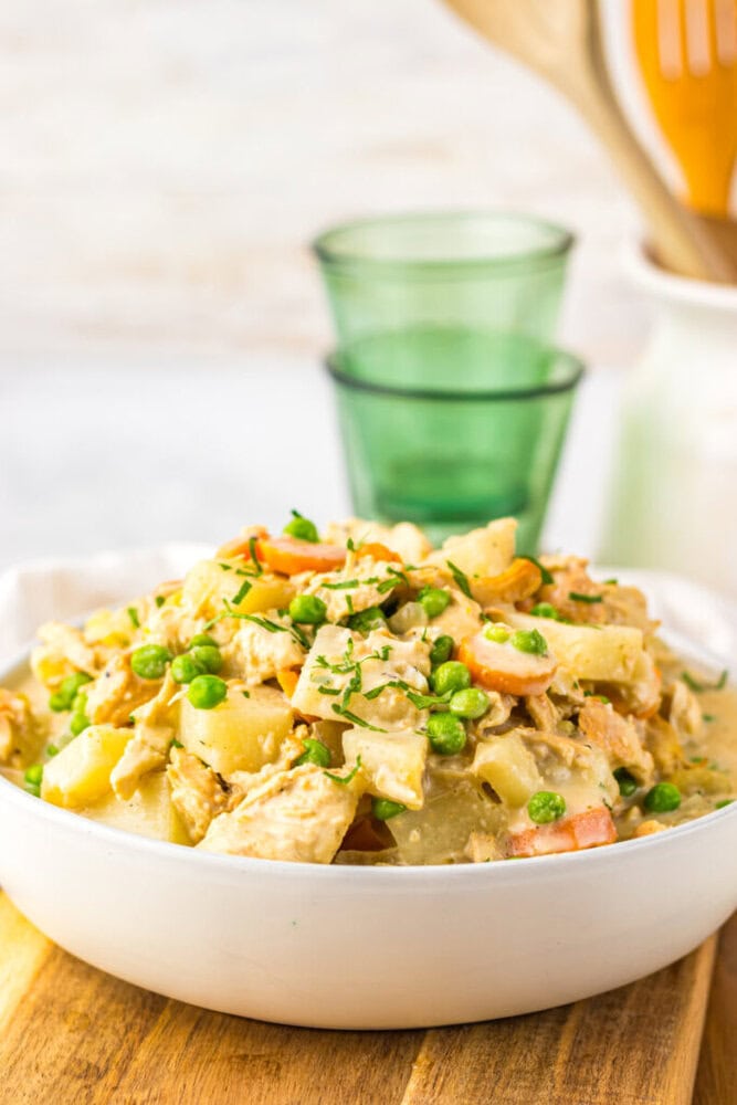 A bowl of creamy chicken stew with potatoes, peas, and carrots sits on a wooden surface with green glasses in the background.