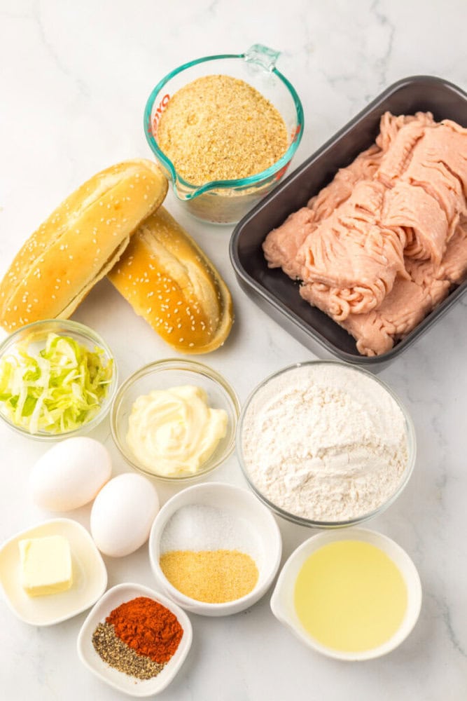 Ingredients for a recipe arranged on a white surface, including ground meat, bread rolls, breadcrumbs, lettuce, mayonnaise, flour, eggs, butter, spices, and lemon juice.