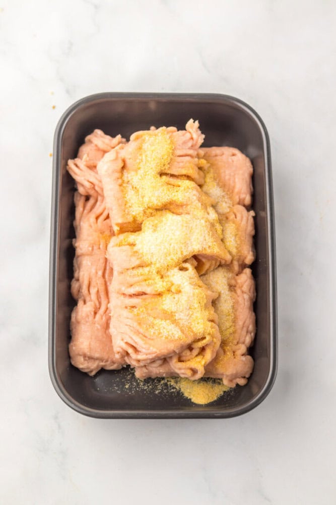 Raw ground meat topped with yellow seasoning is placed in a rectangular metal tray on a white surface.