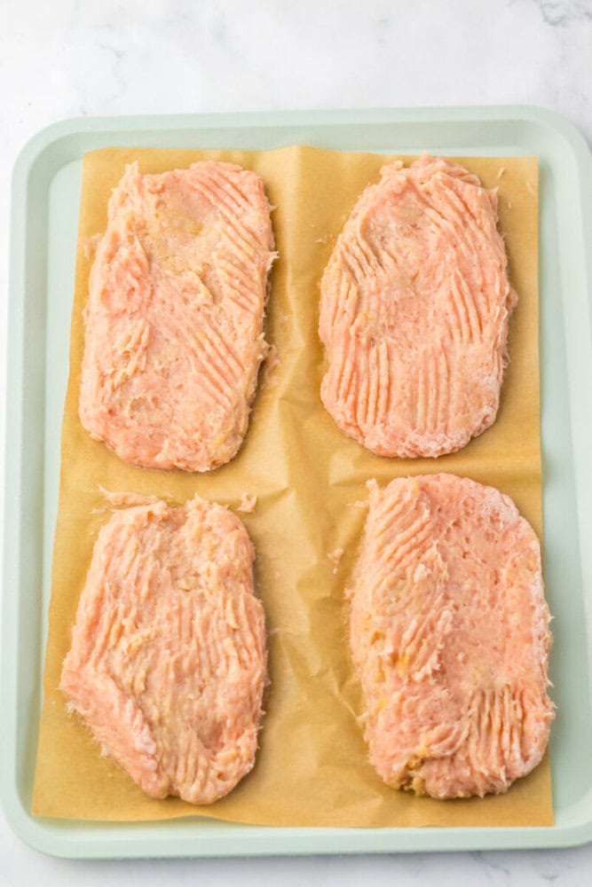 Four raw, rectangular chicken patties are arranged on a sheet of parchment paper on a light green baking tray.