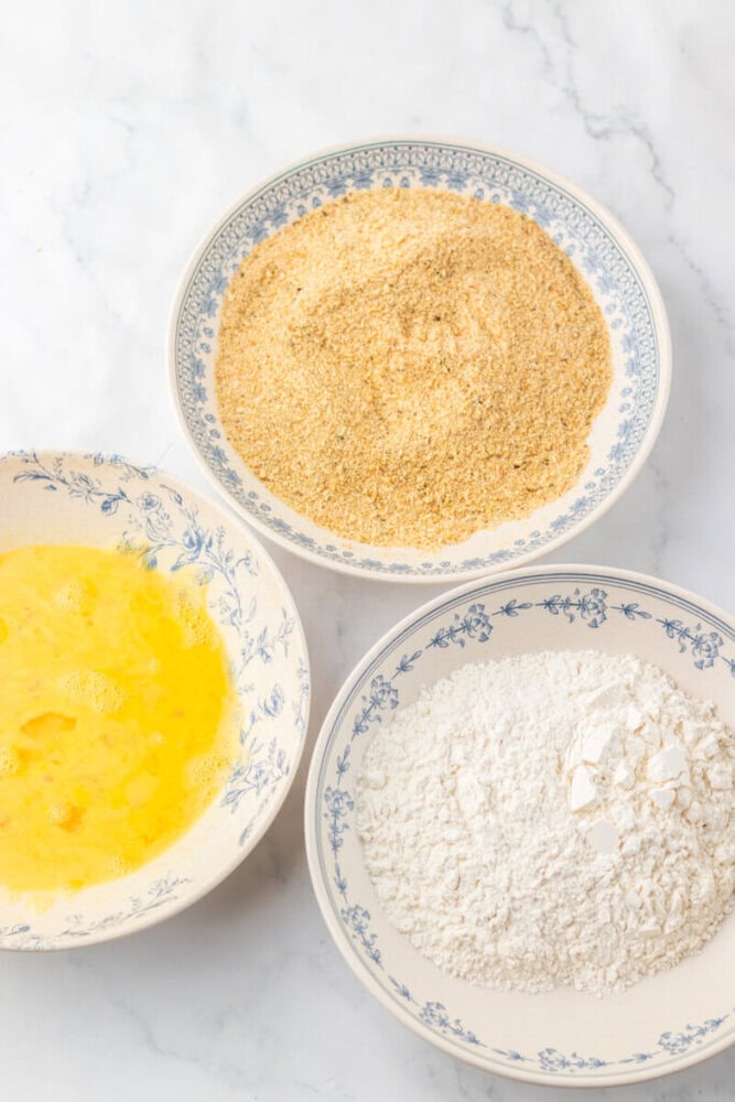 Three shallow bowls contain beaten eggs, breadcrumbs, and flour, arranged on a white marble surface.