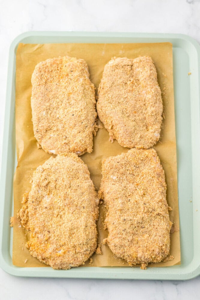 Four breaded cutlets are arranged on a parchment-lined baking tray, ready to be cooked.