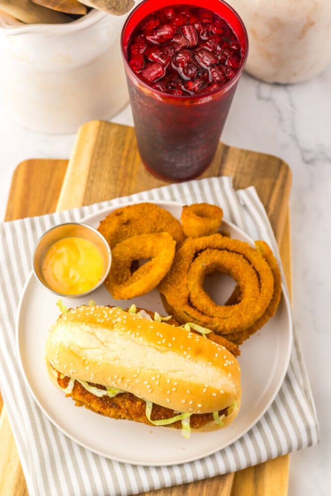 A plate with a breaded chicken sandwich, a side of onion rings, a small cup of dipping sauce, and a red iced beverage on a wooden board with a striped napkin.