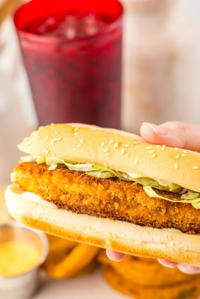 A hand holds a sesame seed bun sandwich with a breaded chicken patty, shredded lettuce, and mayonnaise. A red cup and onion rings are in the background.
