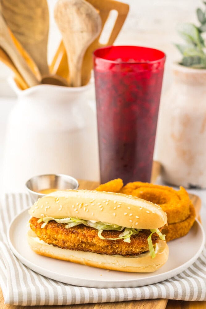 A breaded chicken sandwich with lettuce on a sesame bun is served with onion rings and a cup of iced beverage on a striped napkin.