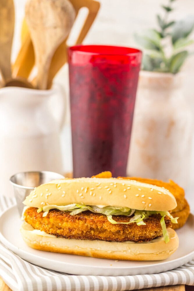 A breaded chicken sandwich with lettuce on a sesame seed bun sits on a white plate next to a metal cup of sauce, with a red drink and kitchen utensils in the background.