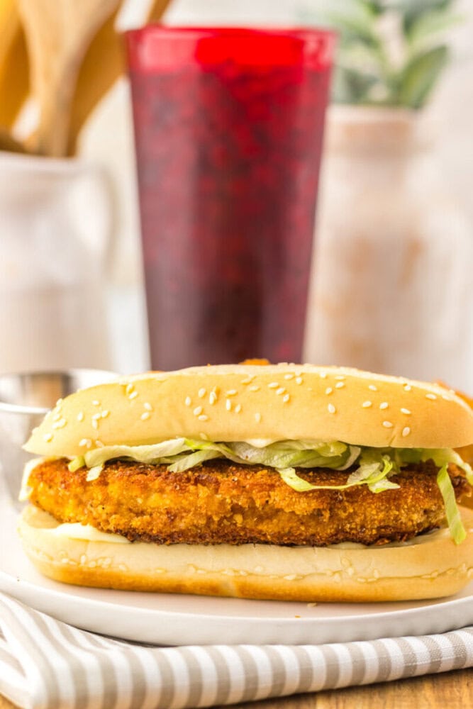 A breaded chicken sandwich with shredded lettuce on a sesame seed bun sits on a plate, with a red glass and potted plant in the blurred background.