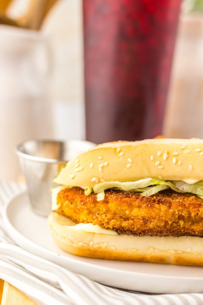 A close-up of a sesame seed sandwich bun filled with a breaded, fried chicken patty, shredded lettuce, and sauce, served on a white plate with a metal condiment cup in the background.