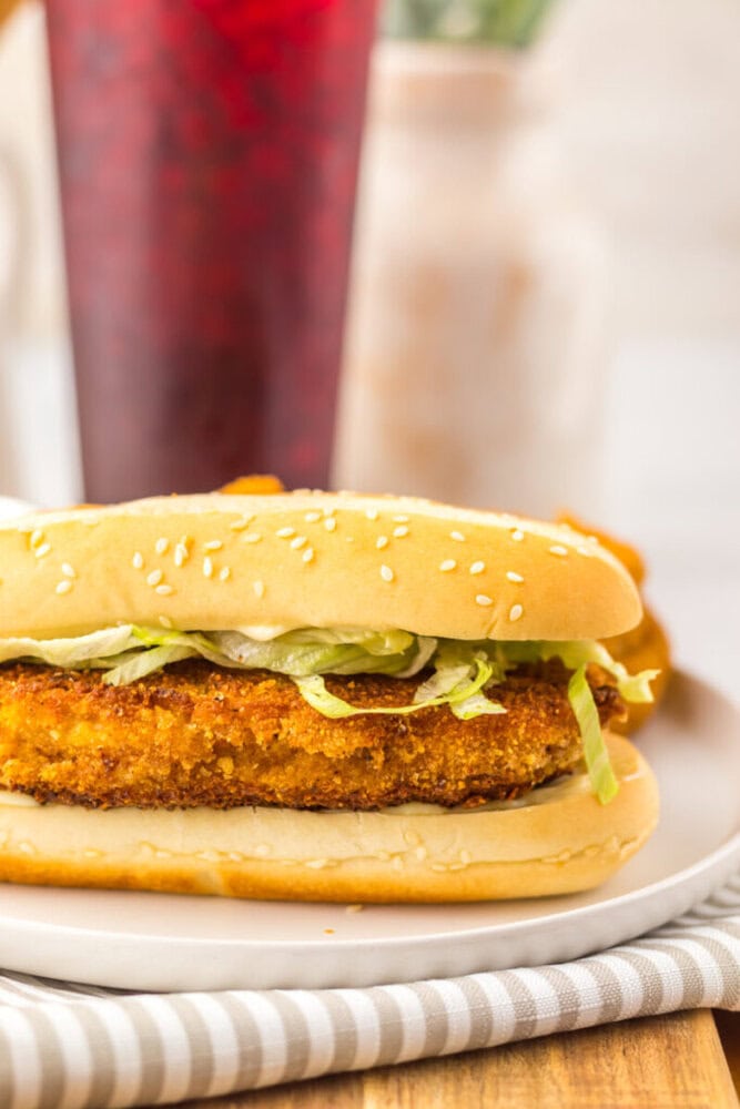 A breaded chicken sandwich with lettuce in a sesame seed bun on a white plate, with a drink and blurred background items.
