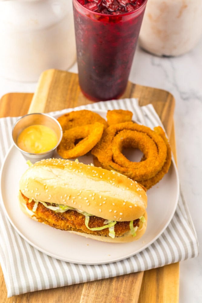 A breaded chicken sandwich with lettuce on a bun, served with onion rings, a side of dipping sauce, and a red iced beverage on a striped napkin.