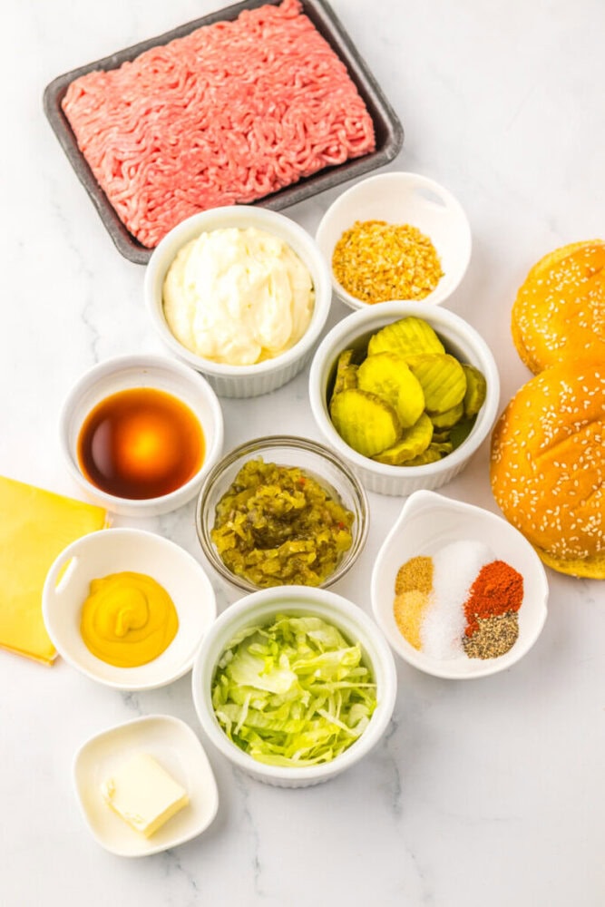 Assorted hamburger ingredients on a white surface, including ground beef, burger buns, cheese, pickles, lettuce, relish, mayonnaise, mustard, seasonings, dried onion, Worcestershire sauce, and butter.