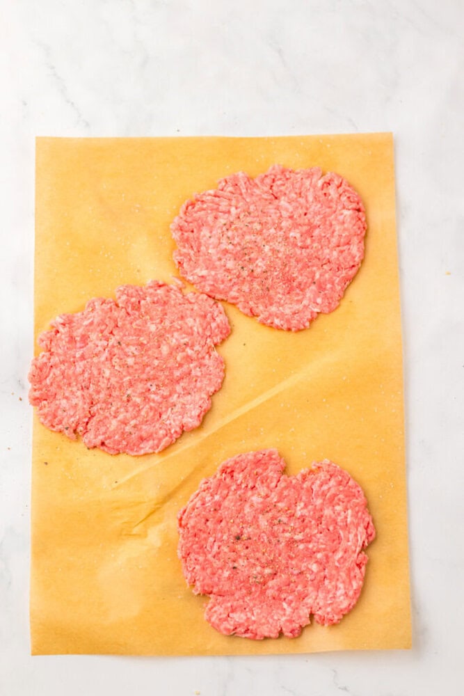 Three raw, seasoned ground beef patties are arranged on a sheet of parchment paper on a white marble surface.