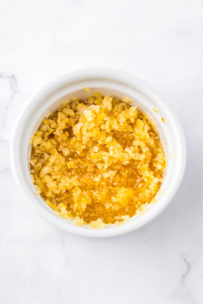 A white ramekin filled with minced garlic in oil, placed on a white marble surface.