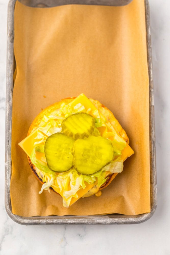 An open-faced cheeseburger with shredded lettuce, cheese, and three pickle slices on a bun, placed on parchment paper in a metal tray.