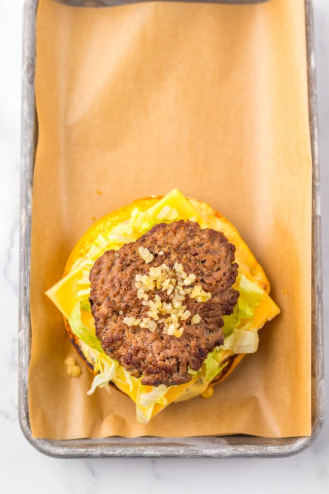 Open hamburger with a beef patty, chopped onions, lettuce, and cheese on a brioche bun, placed on brown parchment paper in a metal tray.