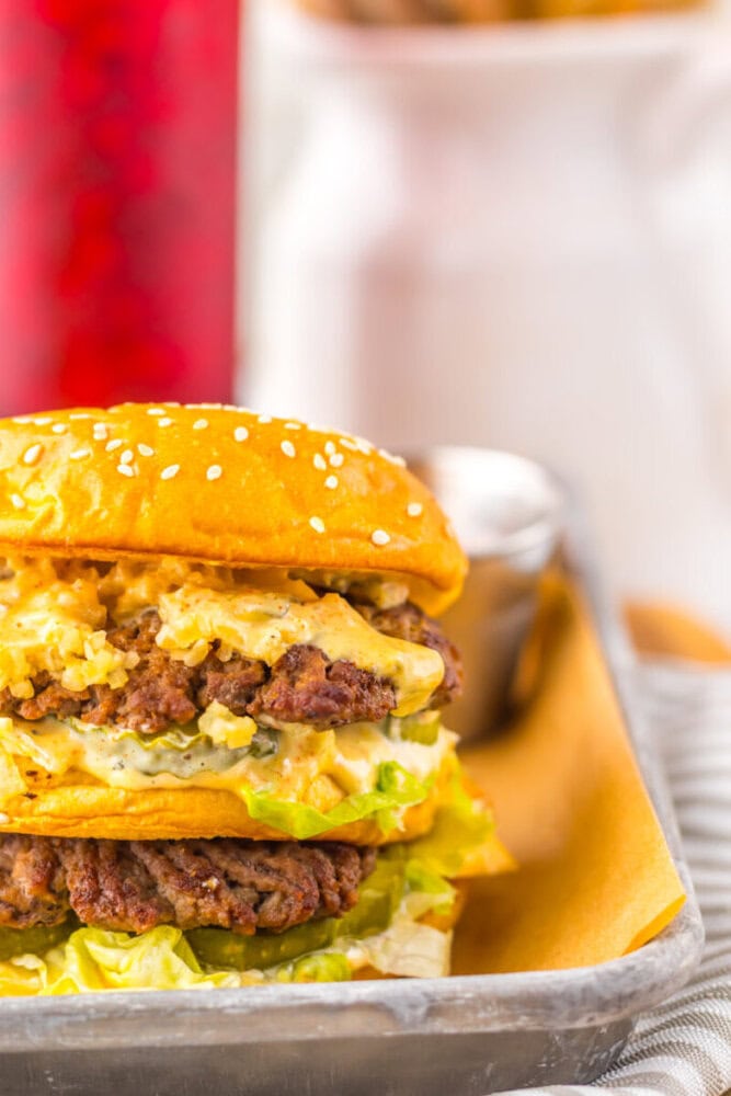 A double cheeseburger with lettuce, sauce, and a sesame seed bun is served in a metal tray lined with parchment paper.