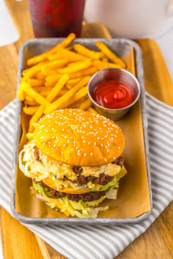 A sesame seed bun double cheeseburger with lettuce and sauce, served with French fries and a cup of ketchup on a tray.