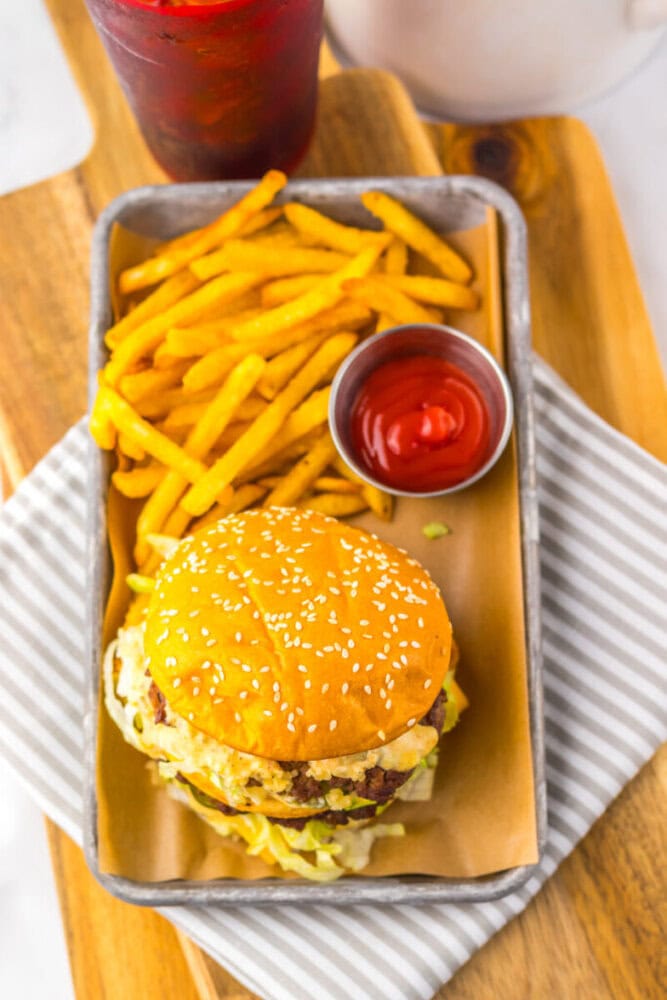 A sesame seed burger bun with a beef patty, lettuce, and cheese served with French fries and a cup of ketchup on a tray, next to a cold drink.