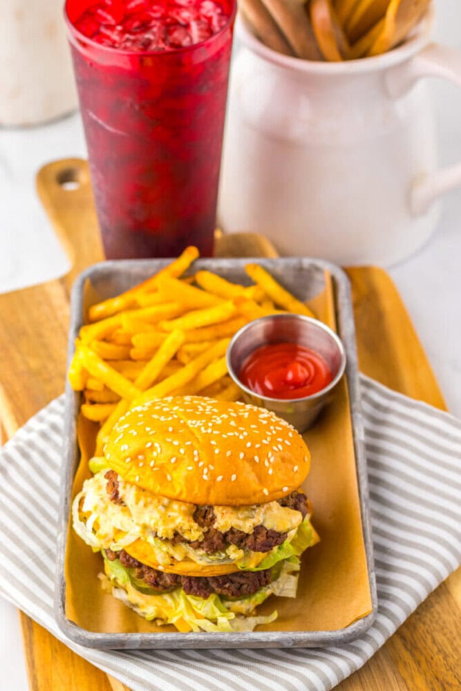 A sesame seed burger with lettuce and cheese, served with a side of French fries and ketchup, and a red iced drink in the background.