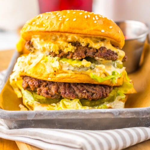 A double cheeseburger with lettuce, pickles, and sauce served on a metal tray with a small cup of dipping sauce in the background.