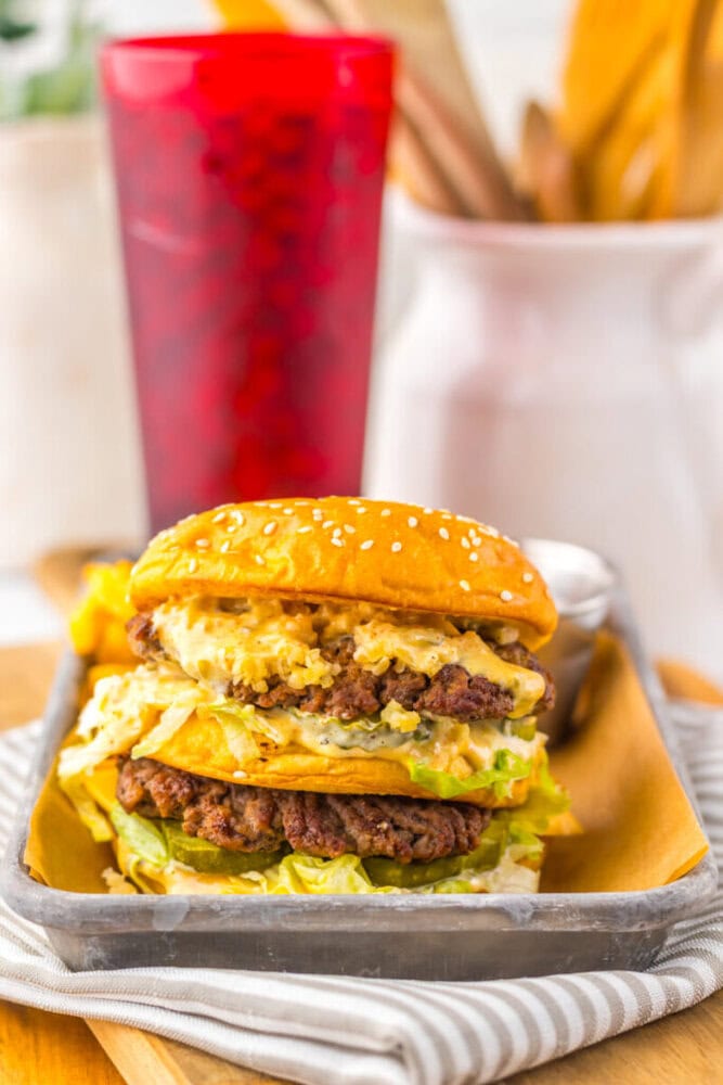 A double cheeseburger with lettuce, sauce, and sesame seed bun is served on a metal tray with a red cup and blurred background items.