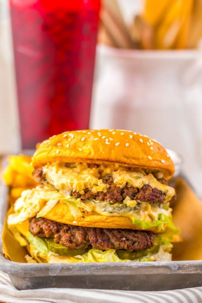 A double cheeseburger with lettuce, sauce, and sesame seed bun sits on a tray with fries. A red beverage is blurred in the background.