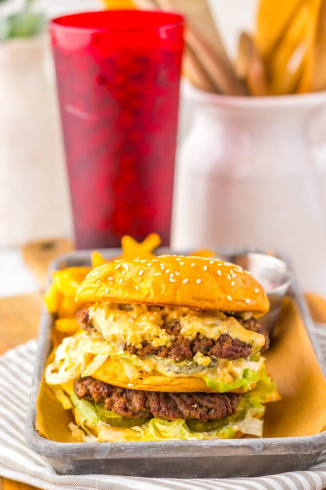 A double cheeseburger with lettuce, sauce, and sesame seed bun is served on a tray with fries, a dipping sauce, and a red cup in the background.