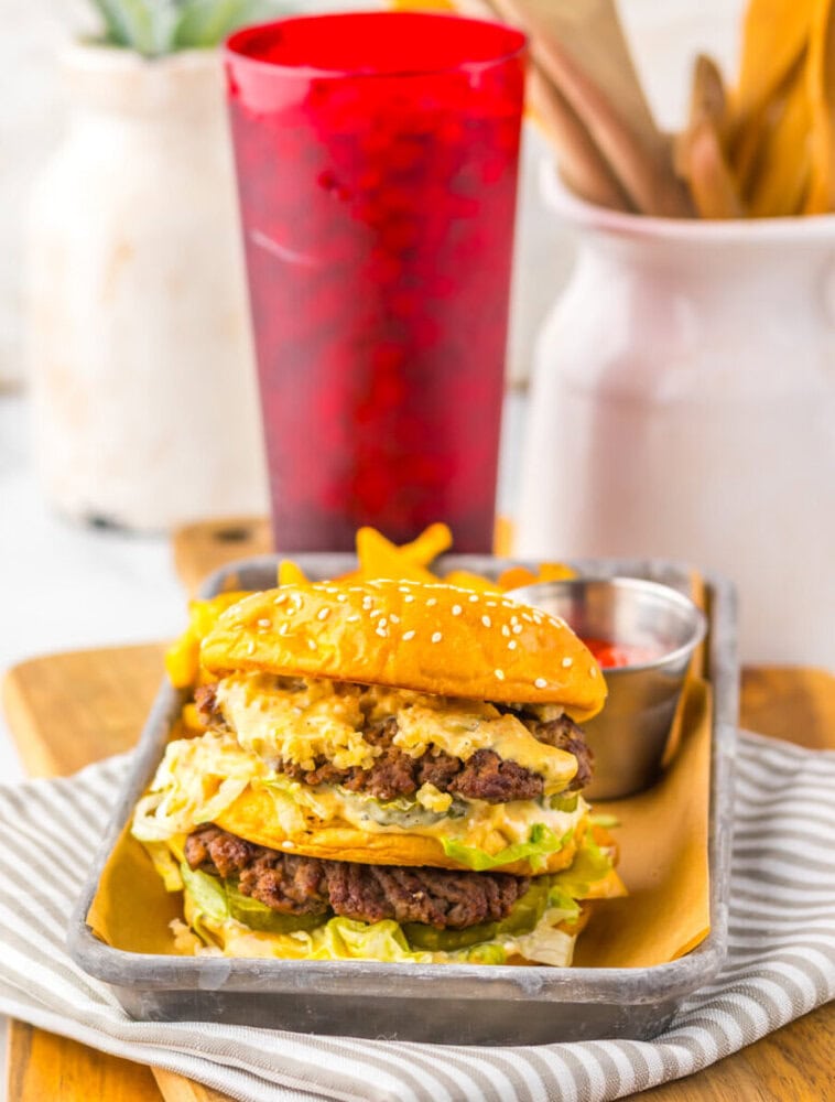 A double cheeseburger with lettuce and sauce on a metal tray, served with a small cup of ketchup and a red drink cup in the background.